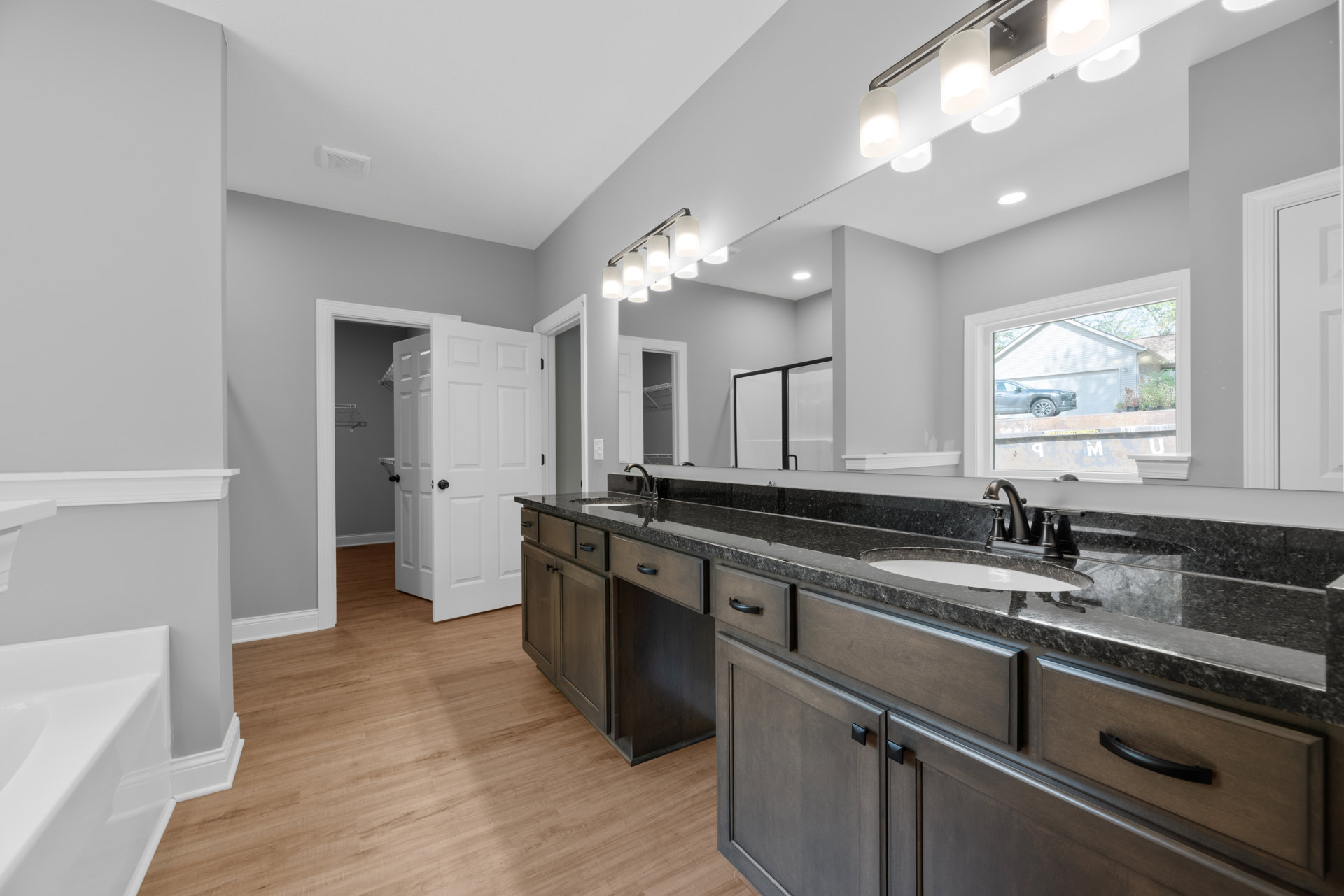 Bathroom with double vanity and wide mirror, white bathtub in corner, wood flooring, window view of parked car, close-up of modern light fixture