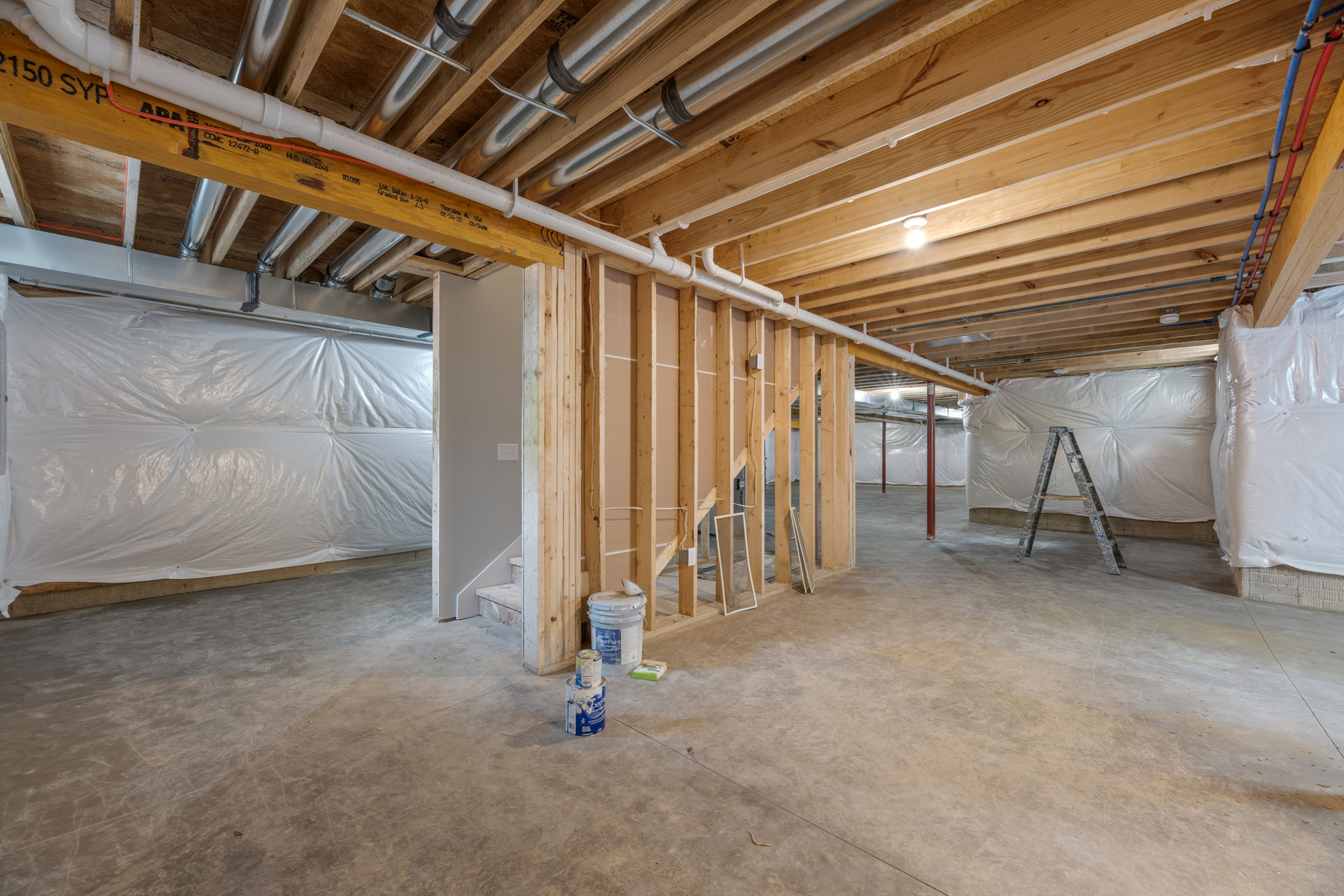 Unfinished basement with exposed beams, plaster walls, two ladders, paint cans, and white plastic sheeting covering part of the wall.