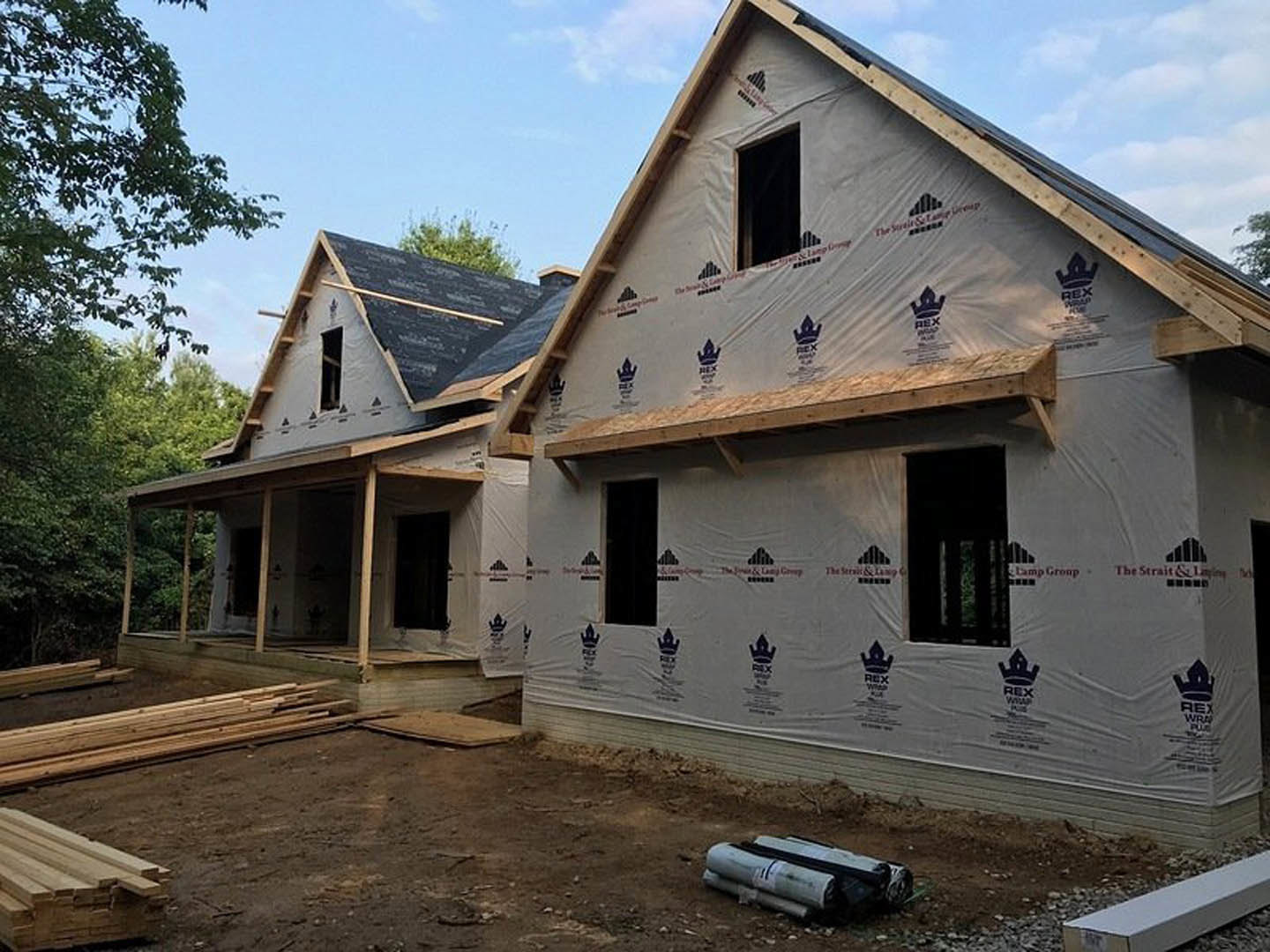 Framed house under construction with exposed wood beams, partially installed windows, stacks of lumber and newspapers on dirt ground, surrounded by trees under clear blue sky