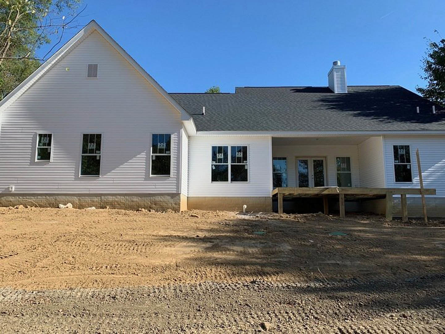 White custom home with multiple windows, dark roof, wooden deck, and dirt yard with tire tracks under clear blue sky