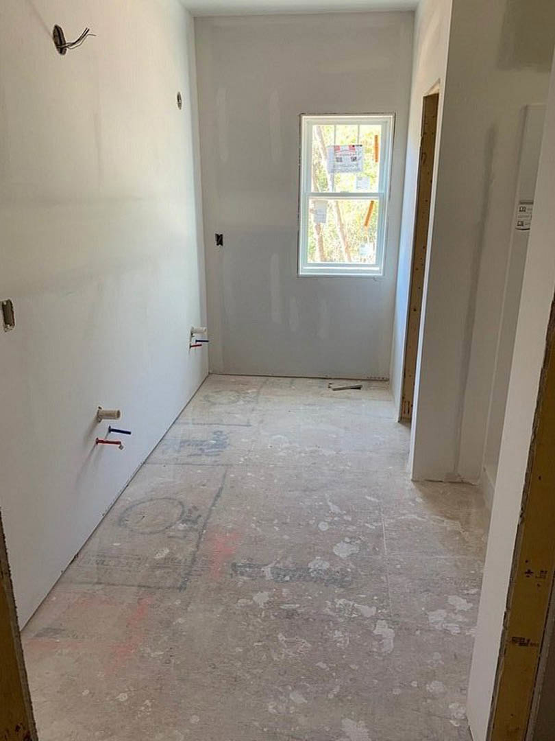 Hallway with light-colored plaster walls, wood flooring, large window, and white ceiling; window features a small sign attached, and wall displays several holes and mounted toilet