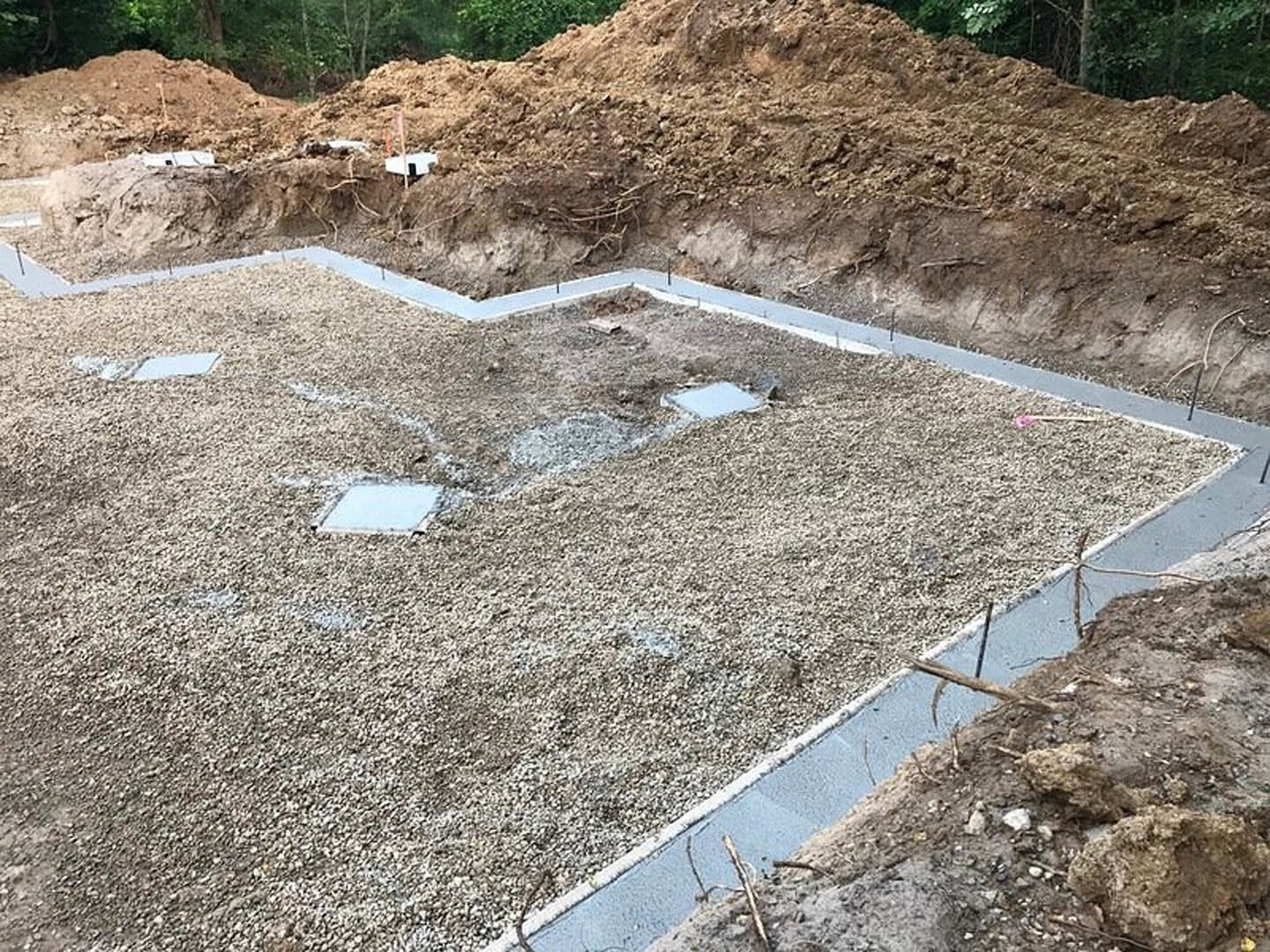 Concrete slab foundation bordered by gravel and dirt, set outdoors under a clear blue sky with nearby curb and sparse vegetation.