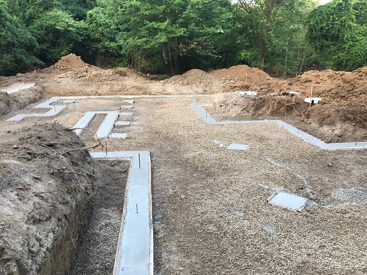 Dirt and gravel piles at a residential construction site bordered by green-leaved trees, exposed soil, and foundation work visible