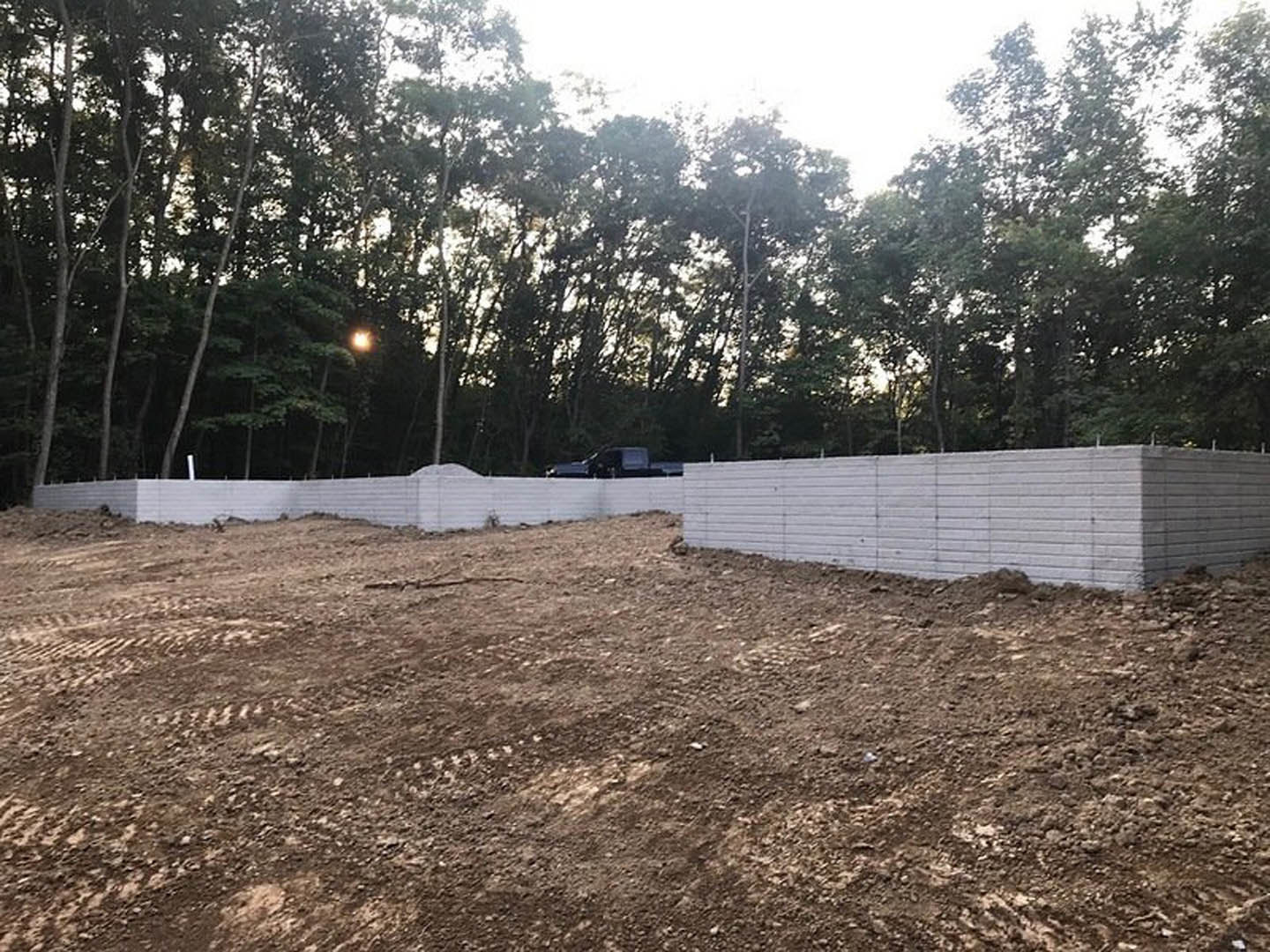 Dirt field bordered by a white brick wall, mature green trees in the background, and a parked truck near the fence