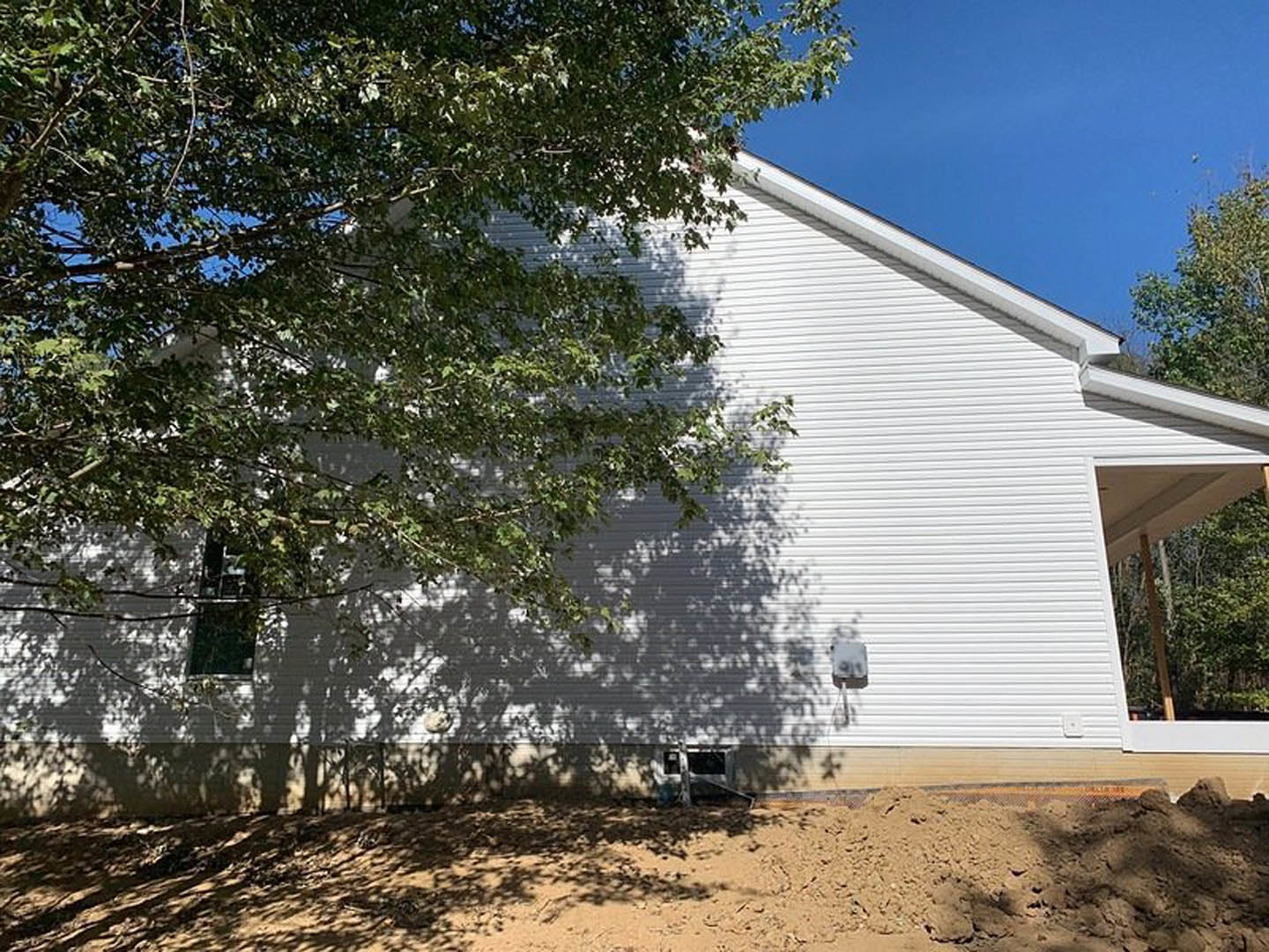 White siding house with white porch and roof, tree in backyard casting shadow on lawn, windows visible, pile of dirt near foundation.