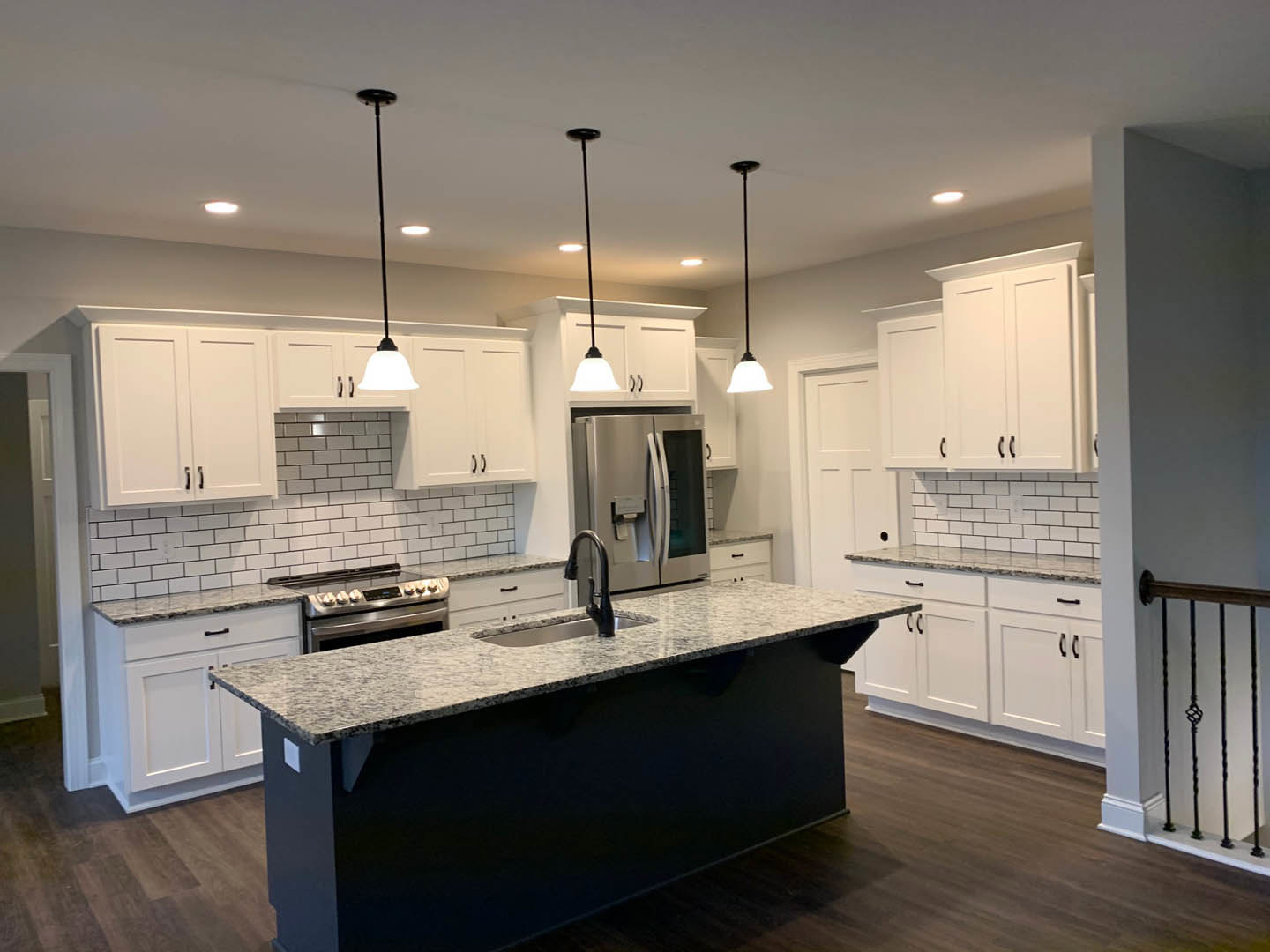 White shaker cabinets, black island with waterfall countertop, stainless steel appliances, farmhouse sink, light wood flooring, pendant lights, and metal railing visible in