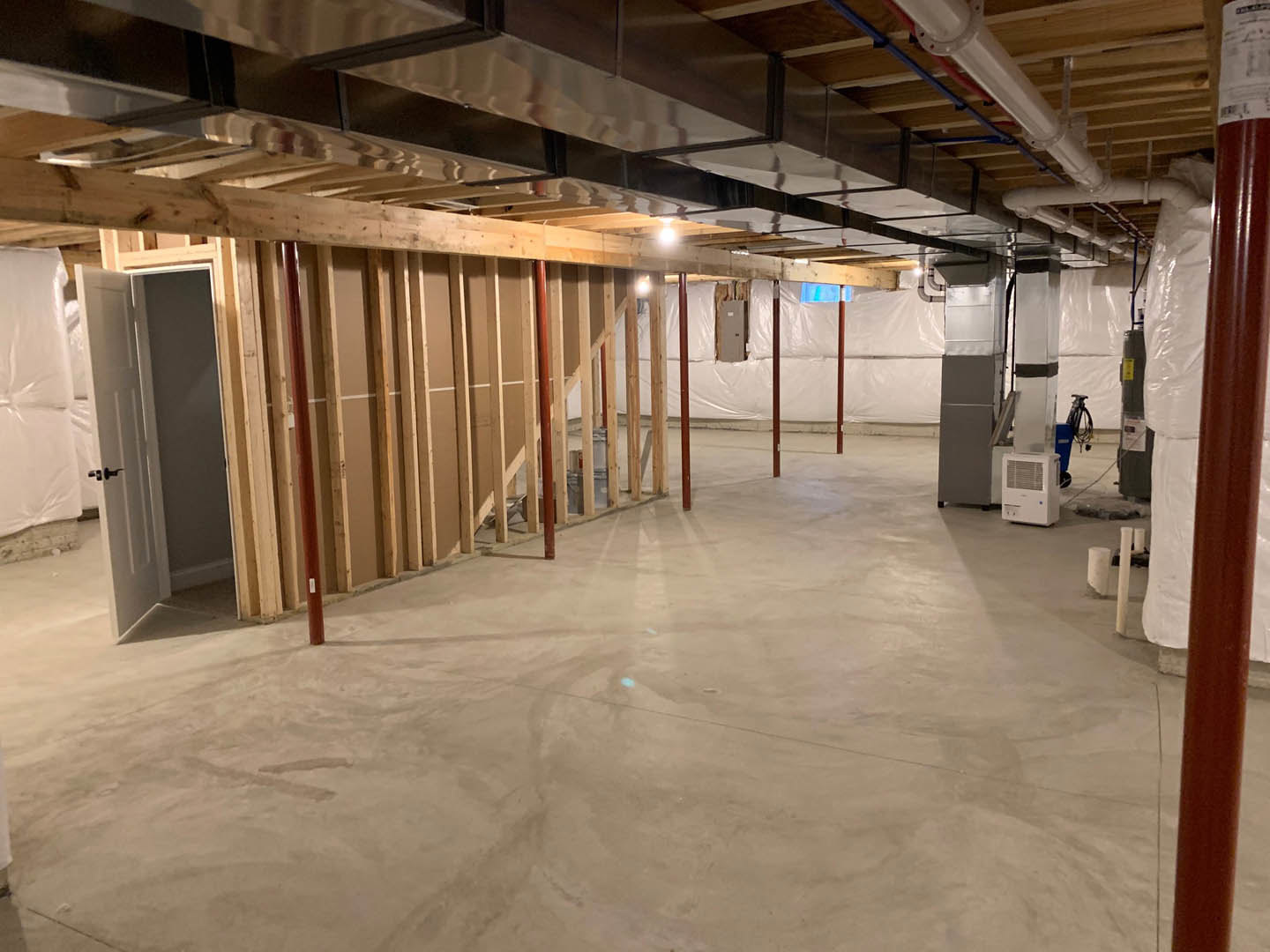 Basement room with exposed concrete floor, white walls, ceiling beams, metal support pole, and a vented rectangular fixture.