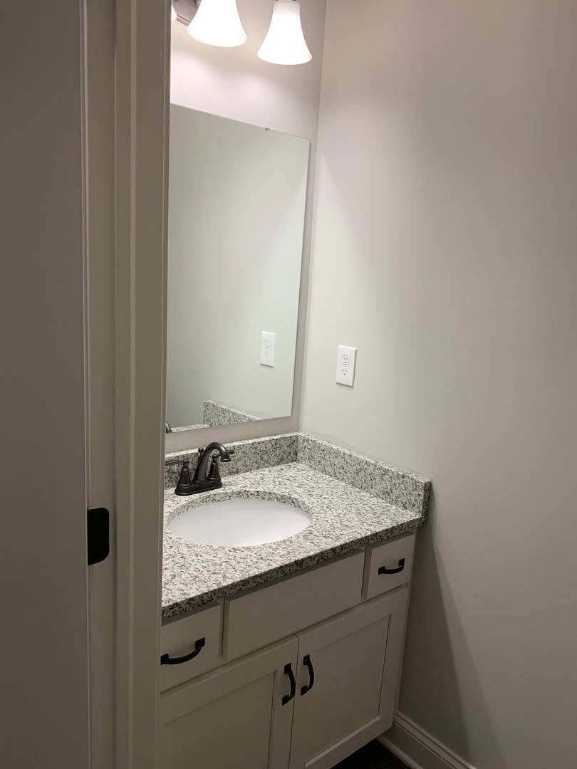 Modern bathroom with white tile walls, rectangular mirror above a sleek sink, chrome faucet, black handle on white vanity drawer, and minimalist light fixture.