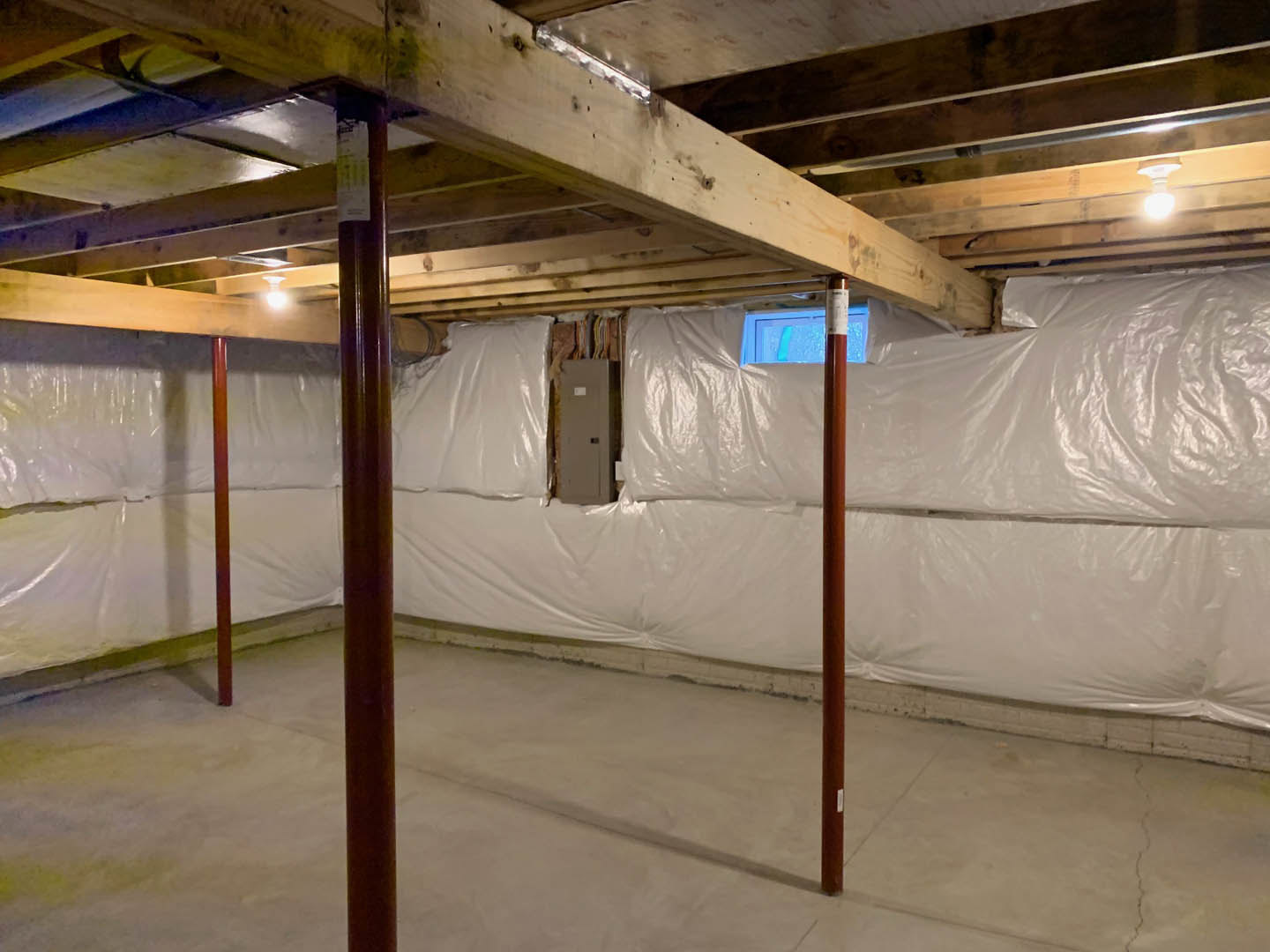 Basement with white plastic covering on concrete floor, exposed beams and walls, white door with sign, close-up of support pole, overhead lighting.