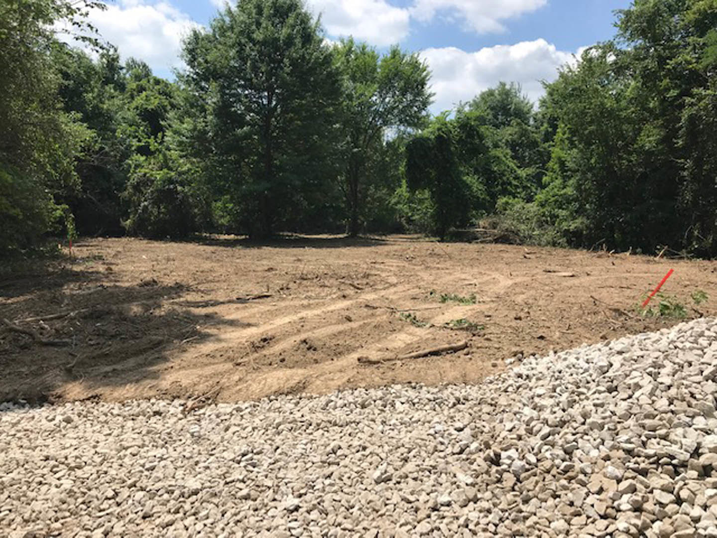 Dirt field with scattered rocks and rubble, bordered by a group of trees under a partly cloudy blue sky