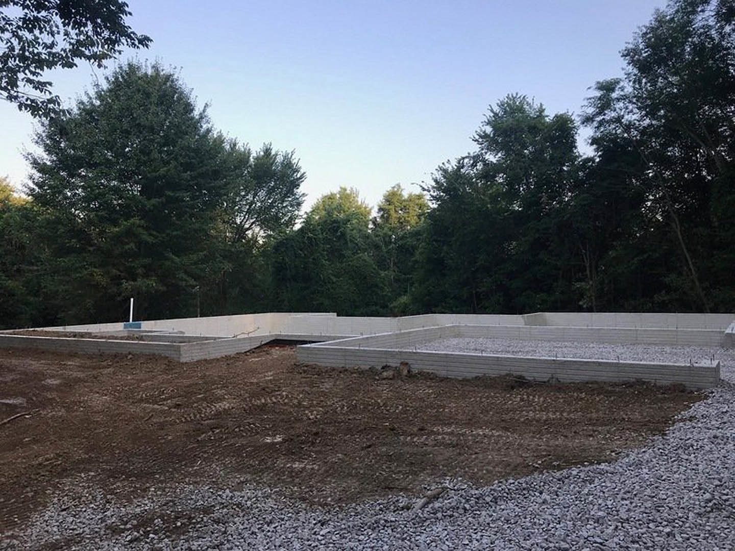 Concrete foundation surrounded by dirt and gravel, patches of snow on the ground, leafy trees in the background under an overcast sky