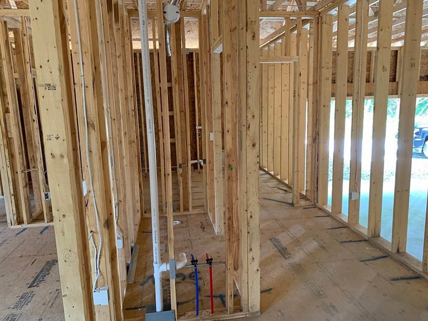 Exposed wood framing with beams and studs, white pipe with red and blue tubes, unfinished interior construction, visible insulation and lumber