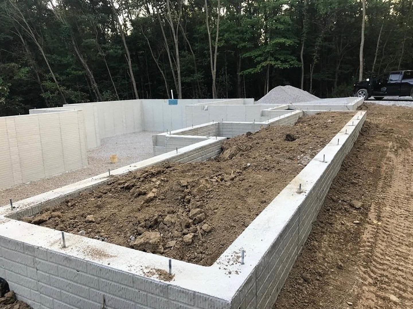 Concrete foundation surrounded by dirt and gravel, wire rods protruding, black truck parked with person in doorway, wooded area with trees in background, pile of white powder near