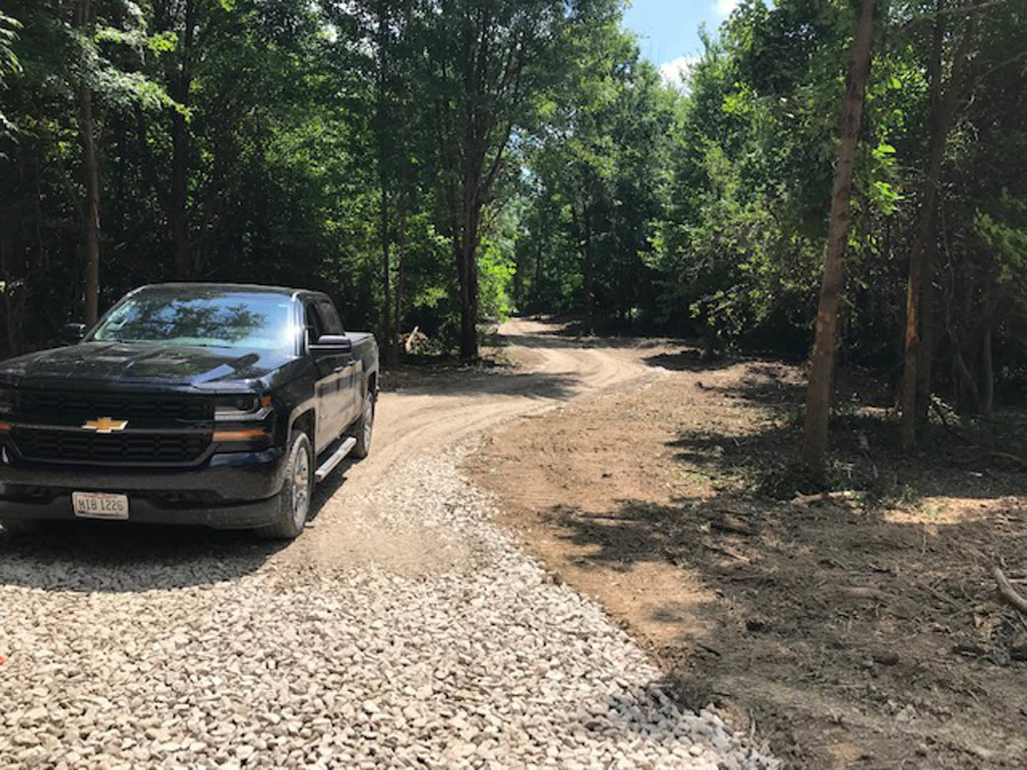 Black pickup truck parked on gravel driveway surrounded by tall green trees under clear blue sky