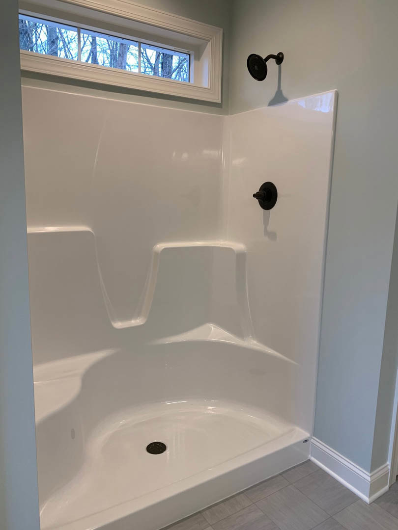 White tile shower with black handle and round shower head, window overlooking trees, close-up of white bathtub
