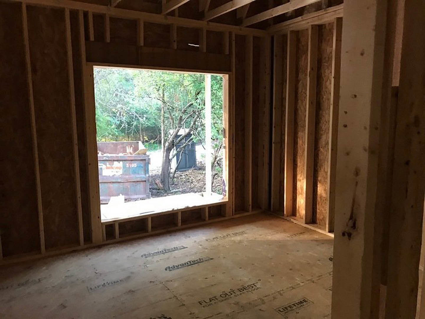 Unfinished room featuring exposed wood framing, large window overlooking trees, plywood floor marked with black text, and a shelf with drilled holes.