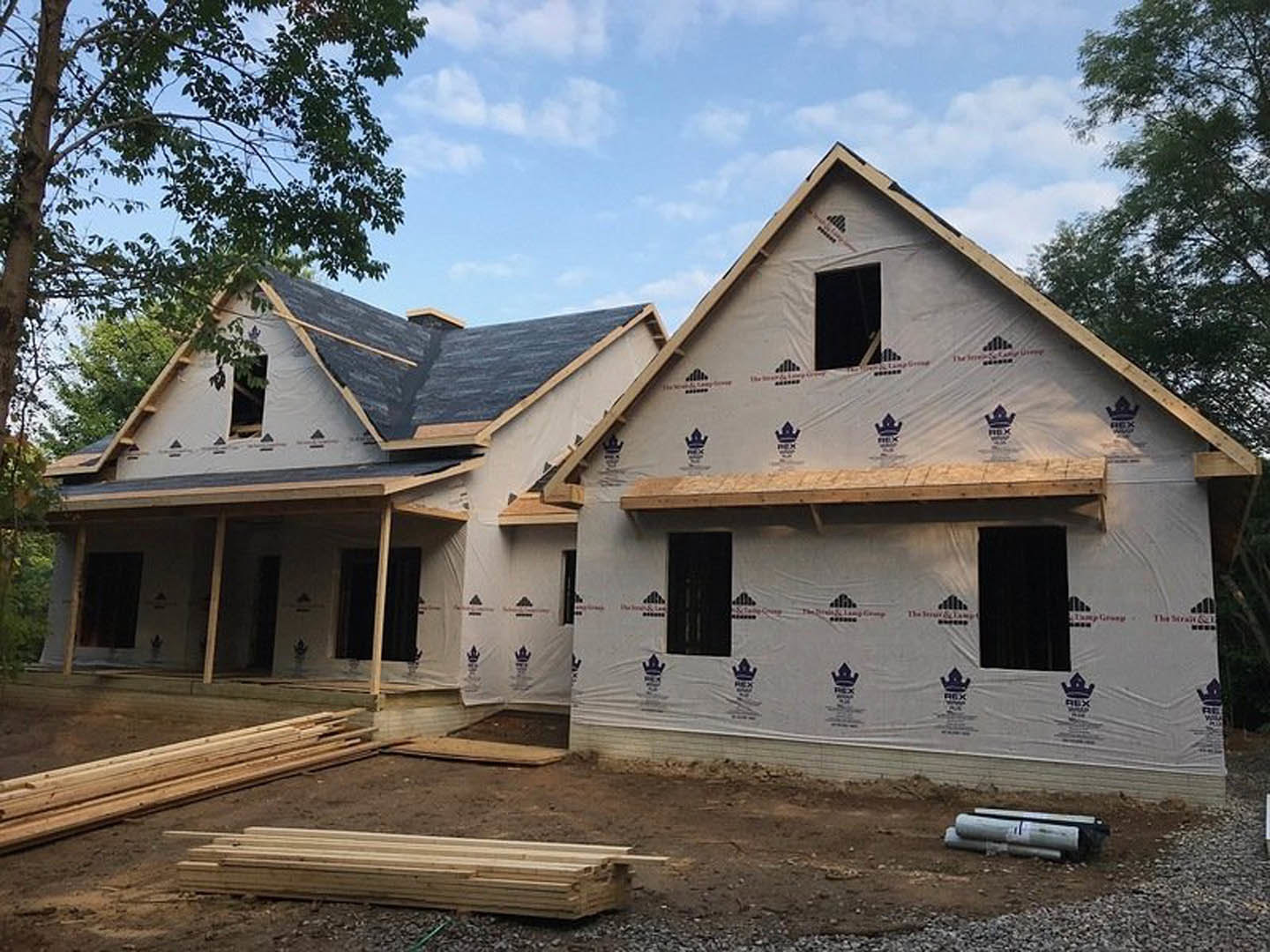 Two-story house under construction with exposed wooden framing, unfinished roof, and stacks of lumber on the ground; cloudy sky and trees in background