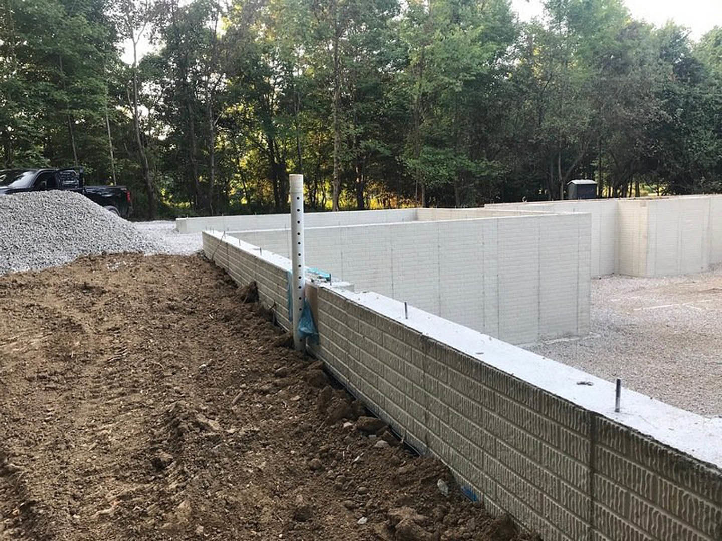 Construction site with a mound of dirt and gravel, partially built white brick foundation wall, trees in background, vehicle parked nearby