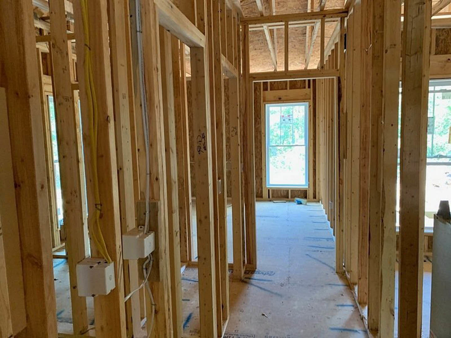 Room under construction with exposed wood framing, visible beams, a window displaying a sign, hallway area, and electrical boxes with yellow and white wires.