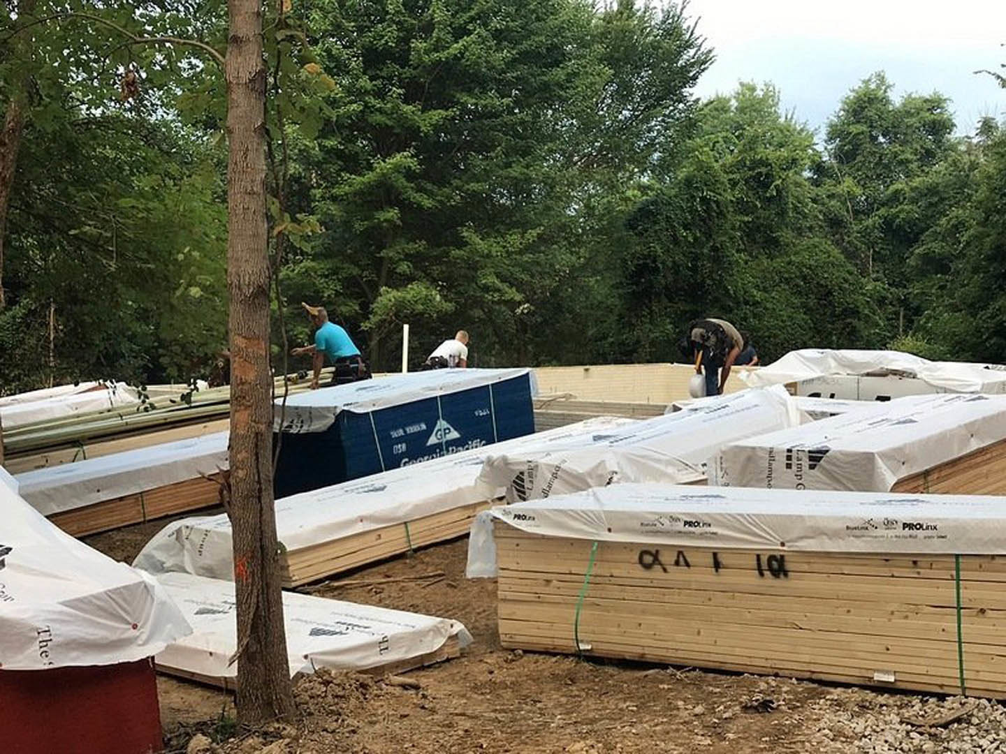 Workers assembling wooden framing and white plastic sheeting on a residential construction site, surrounded by dirt ground, trees, and open sky.