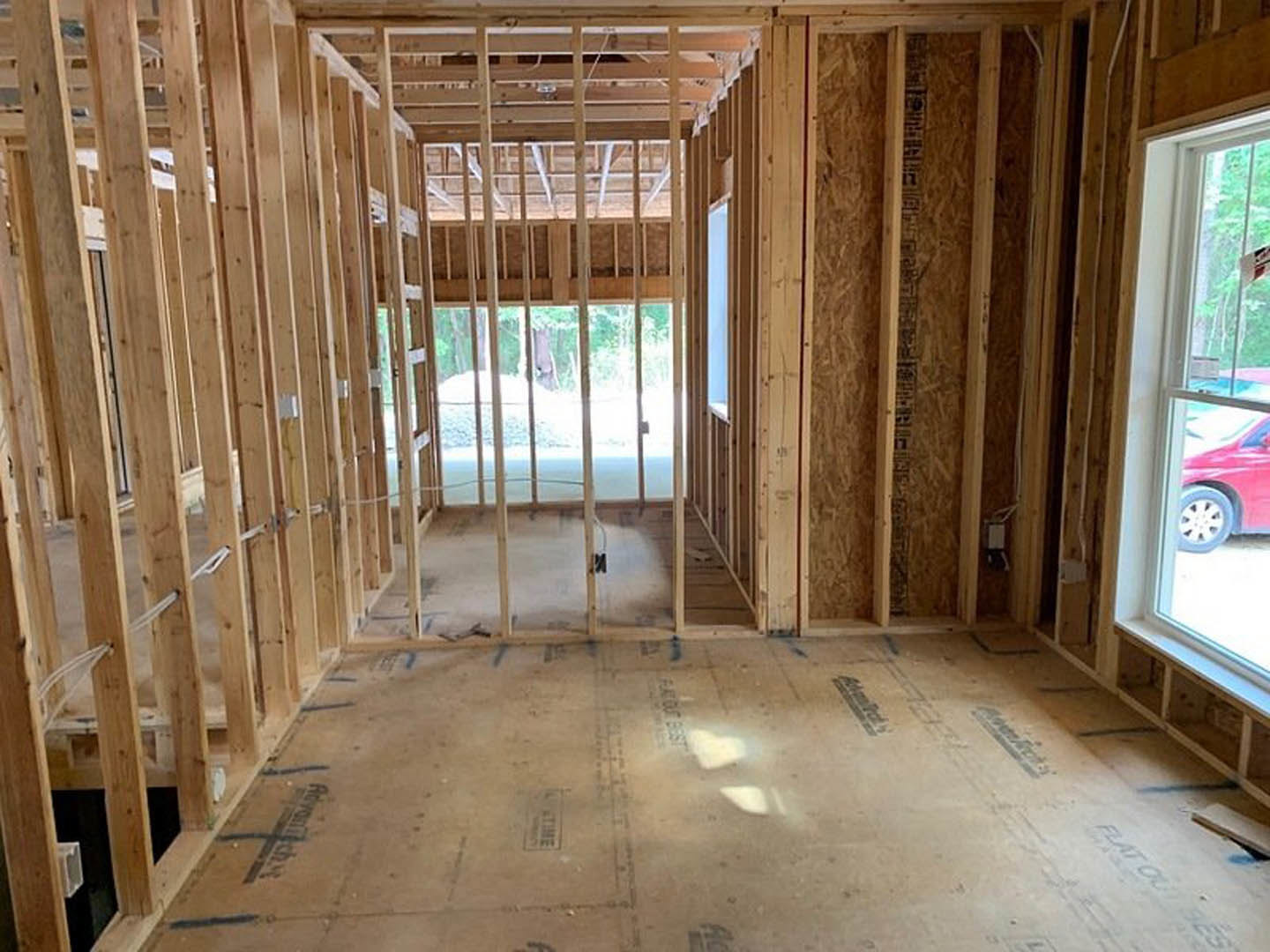 Wood-framed room under construction with exposed beams, unfinished flooring, and a window revealing a red car parked outside