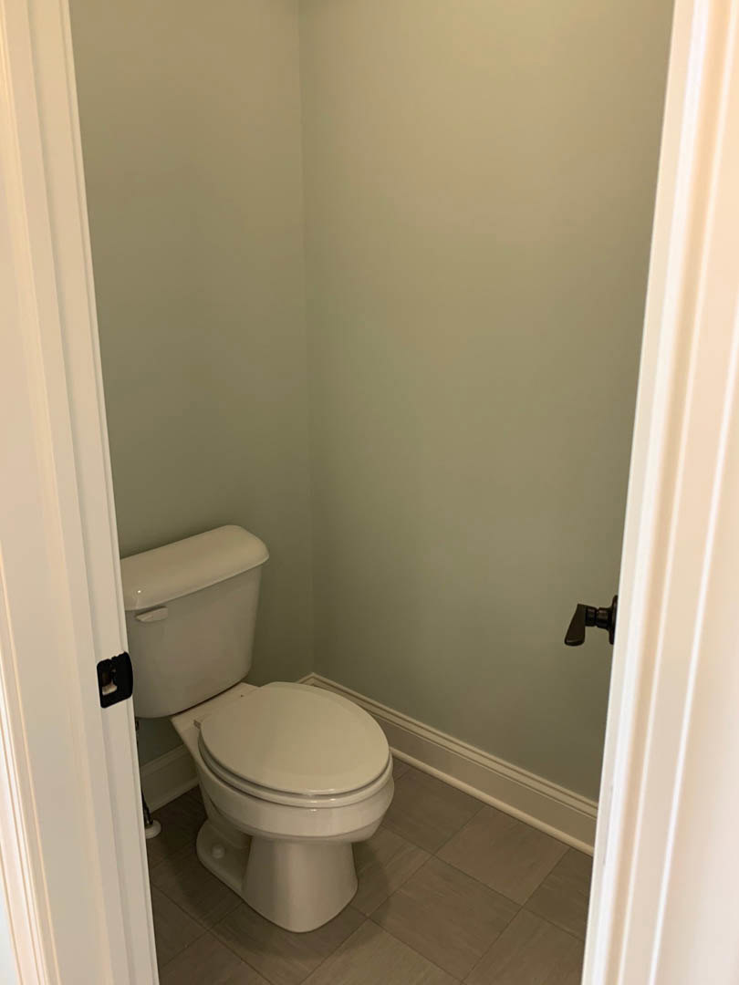 White porcelain toilet with lid up, chrome flush handle on tank, light gray tile floor, white painted wall, partial view of white door