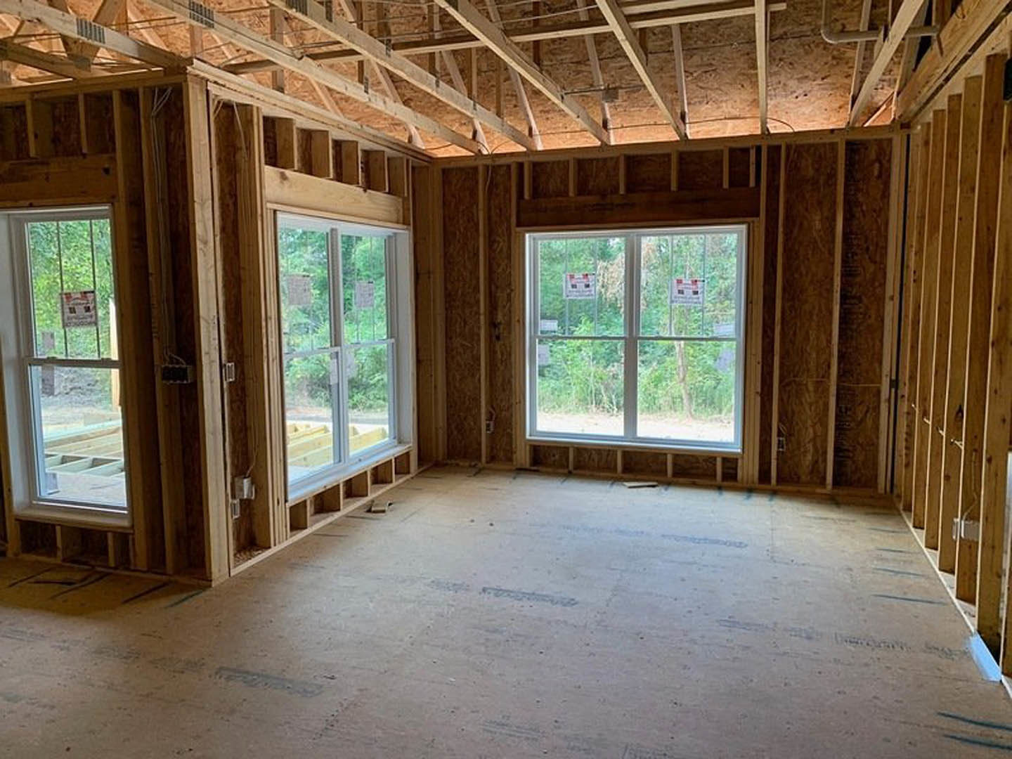 Open room with large windows, exposed wood ceiling beams, light plank flooring, and black electrical outlet with white cord.