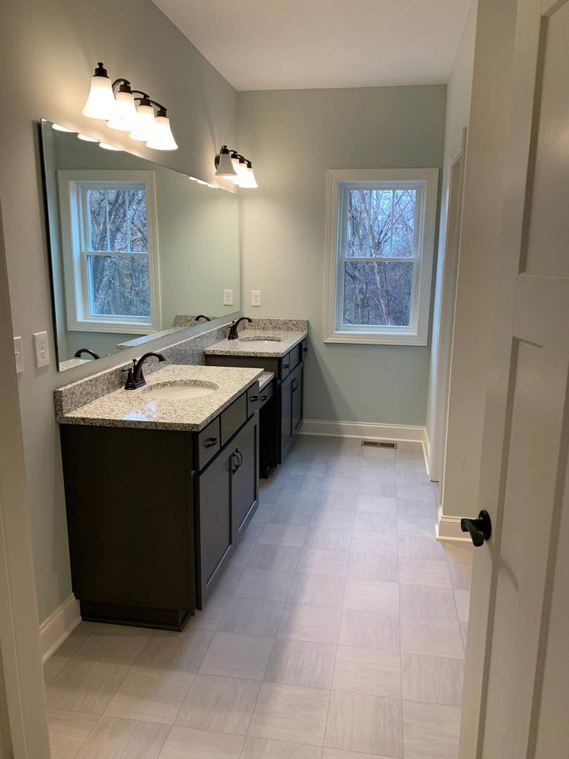 Bathroom with double vanity, rectangular mirror, white sinks, chrome faucets, light gray tile backsplash, window showing leafy tree, white cabinetry, marble countertop, and wood