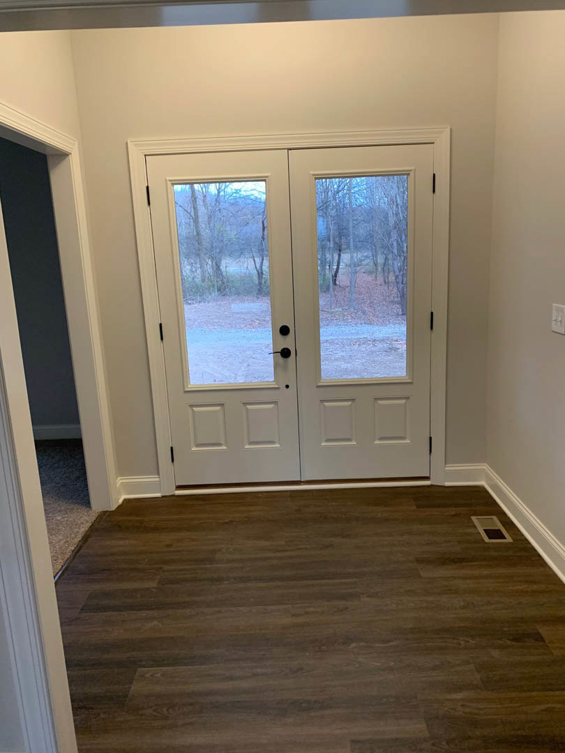 Double glass-paneled doors with black handles set in a white wall, wood flooring, partial view of white cabinetry, and wooded landscape visible through the doors