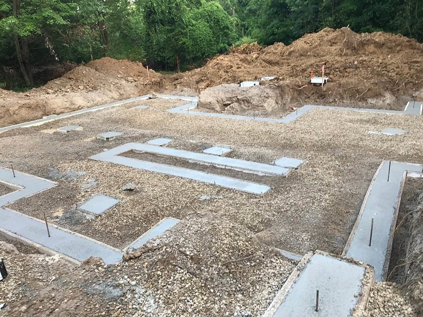 Concrete foundation slab surrounded by dirt piles, scattered construction materials, and trees at a residential building site