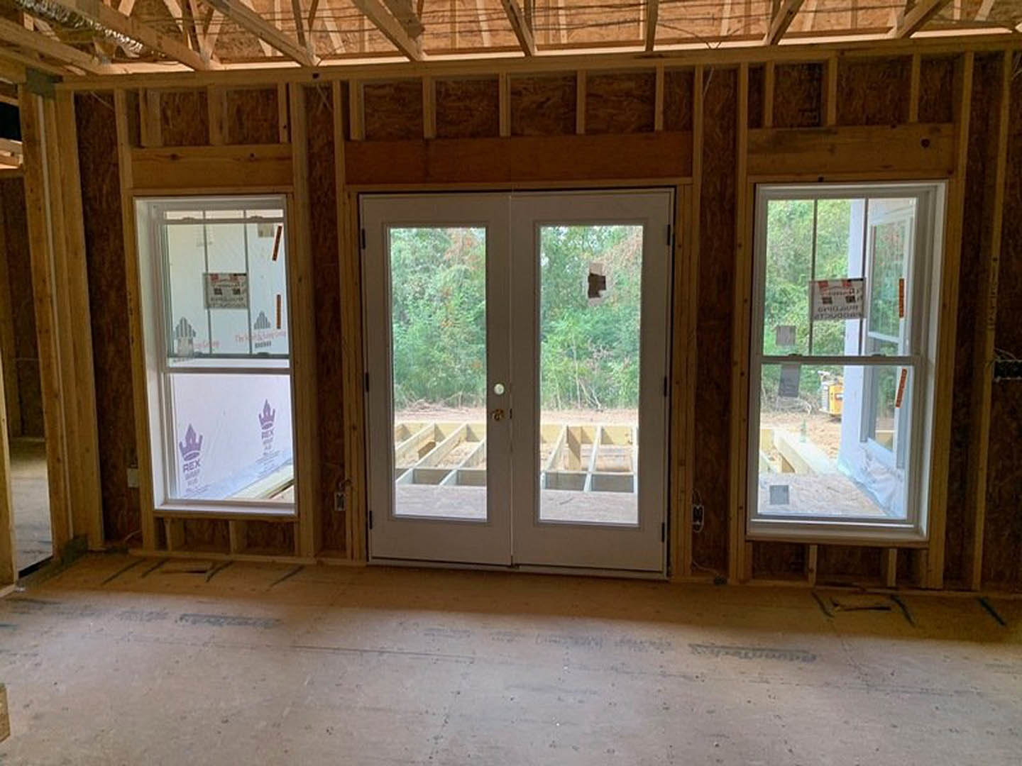 Room with unfinished wood floors, double glass-paneled doors, large windows, and visible construction materials; black and white sign taped to window.