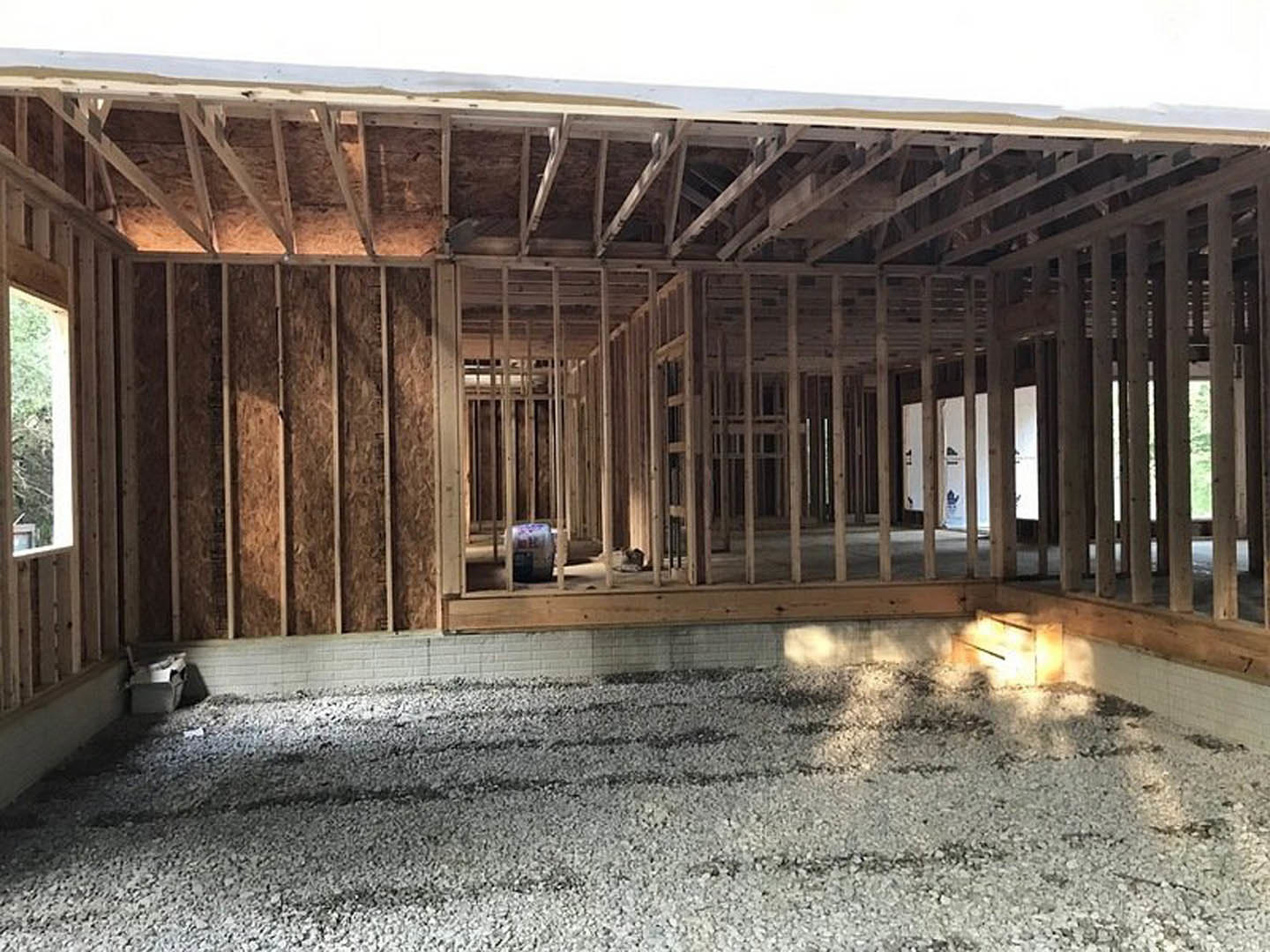 White brick exterior wall and exposed wooden beams of a house under construction, gravel covering the ground, large window framing a tree in the background