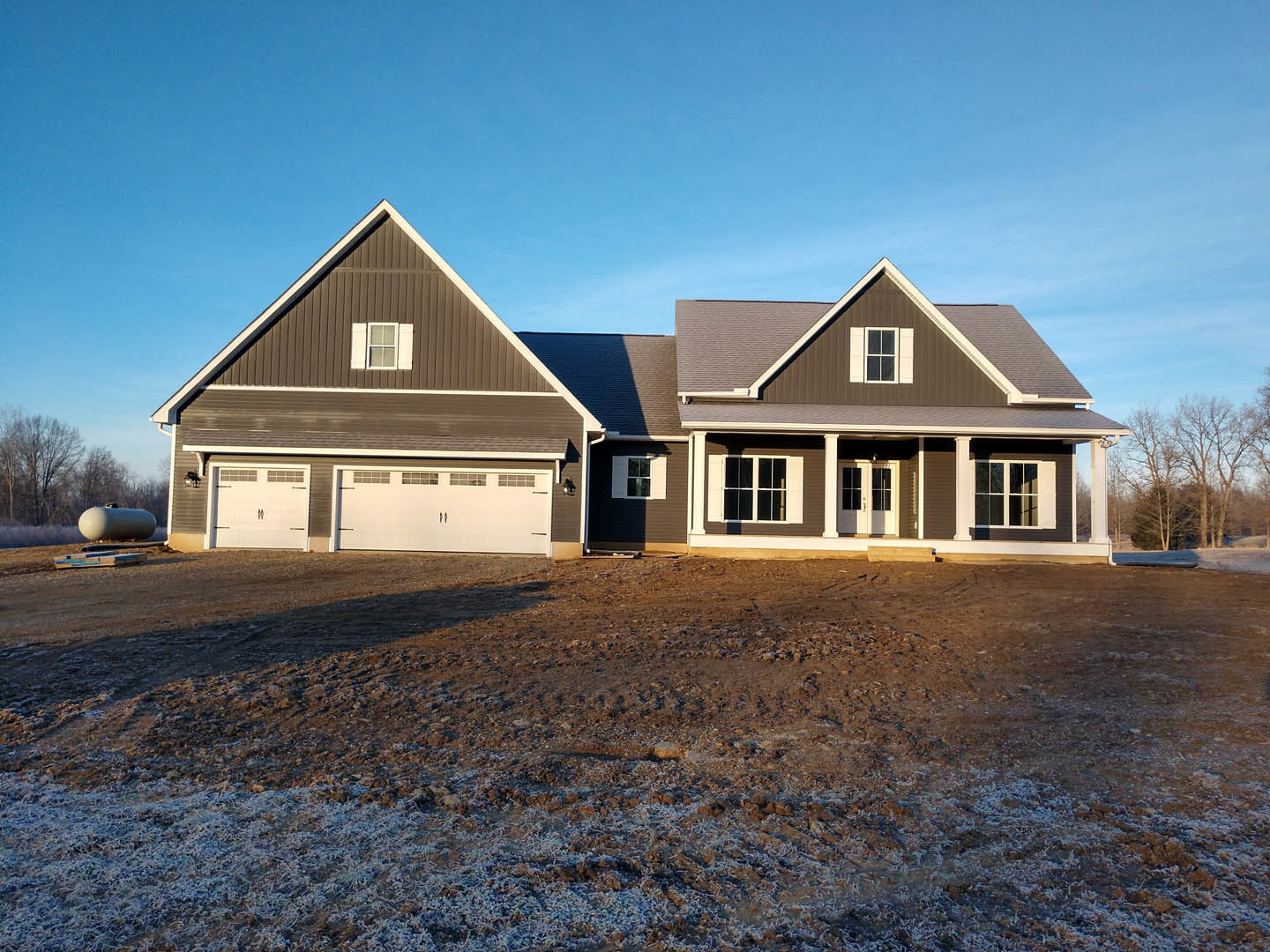 White custom home with attached garage, dirt driveway, white-trimmed windows, and row of trees under clear blue sky