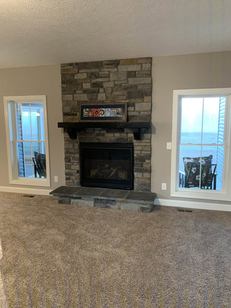 Stone fireplace with black glass door, wood flooring, large windows overlooking ocean, upholstered chair with pillow, white walls