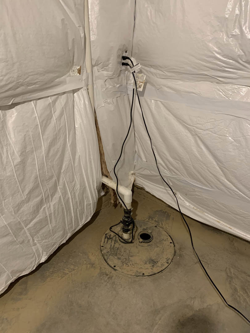 Metal floor drain surrounded by concrete basement floor, white plastic bag with black wire emerging from white pipe nearby, unfinished walls in background.
