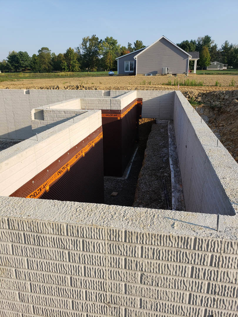Partially built house with exposed concrete and brick walls, roof framing in progress, mature trees and finished home visible in the background