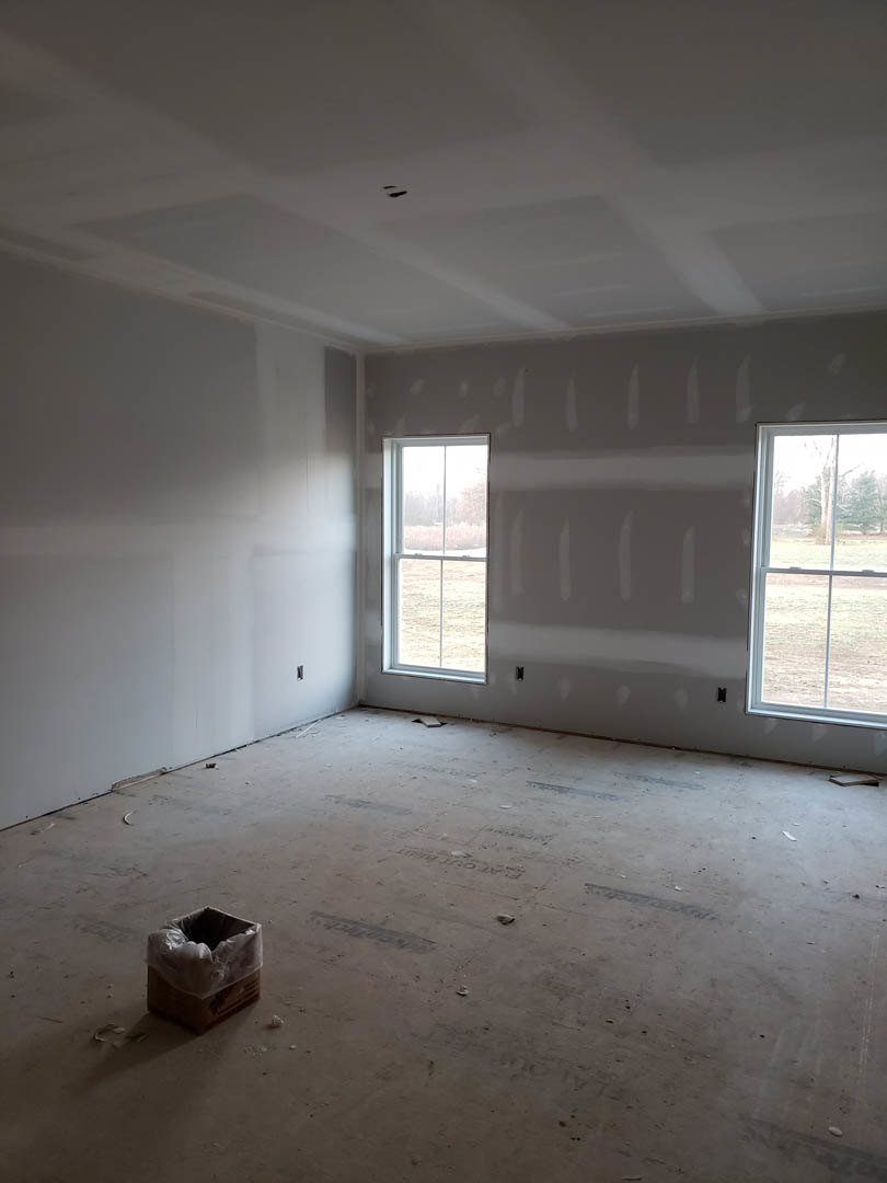 Neutral-toned plaster walls and ceiling, two large windows with views of grassy field, light wood flooring, brown cardboard box with white plastic bag near wall
