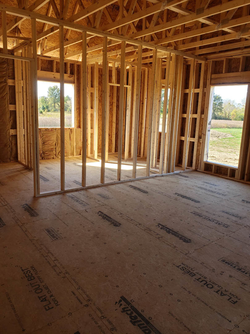 Room under construction with exposed wood framing, ceiling beams, unfinished walls, and a window overlooking a grassy field