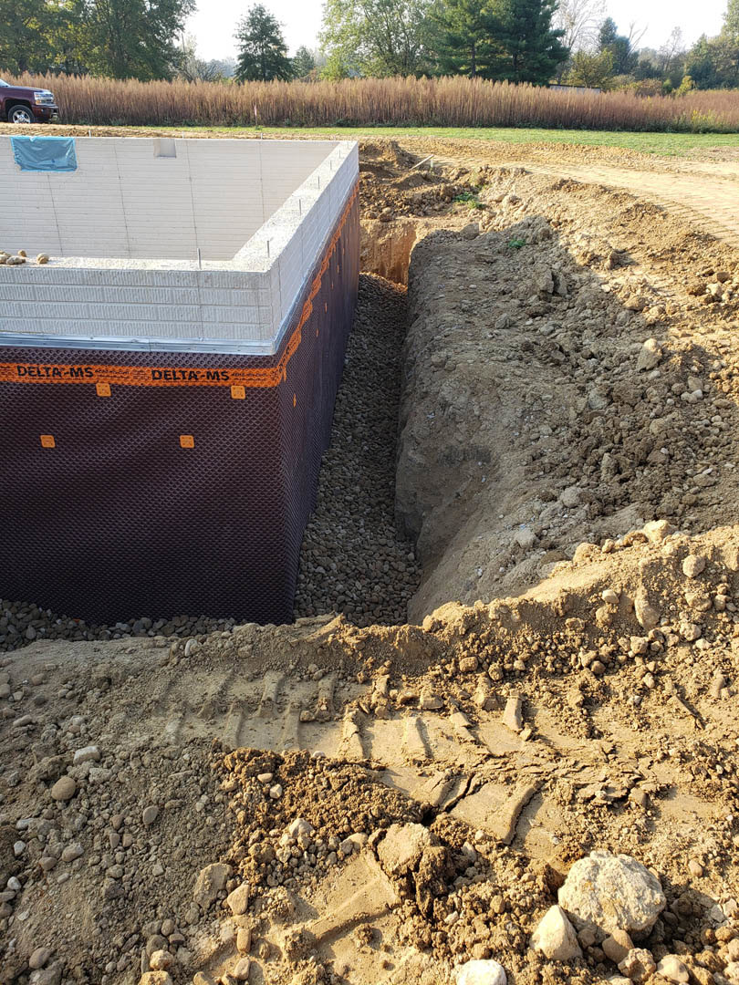 Concrete slab foundation surrounded by exposed soil, tall grass, and trees in the background, with a blue tarp partially covering a white wall.