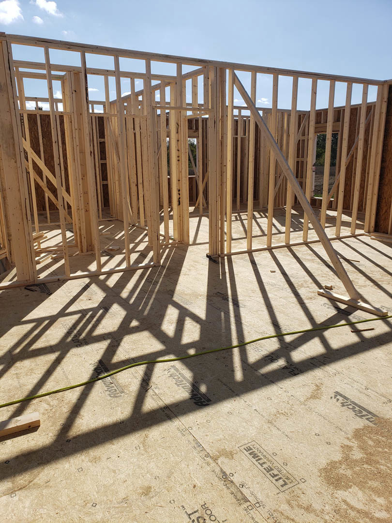 Wooden house frame under construction with exposed beams, green hose on ground, cardboard box and construction sign nearby, cloudy sky in background