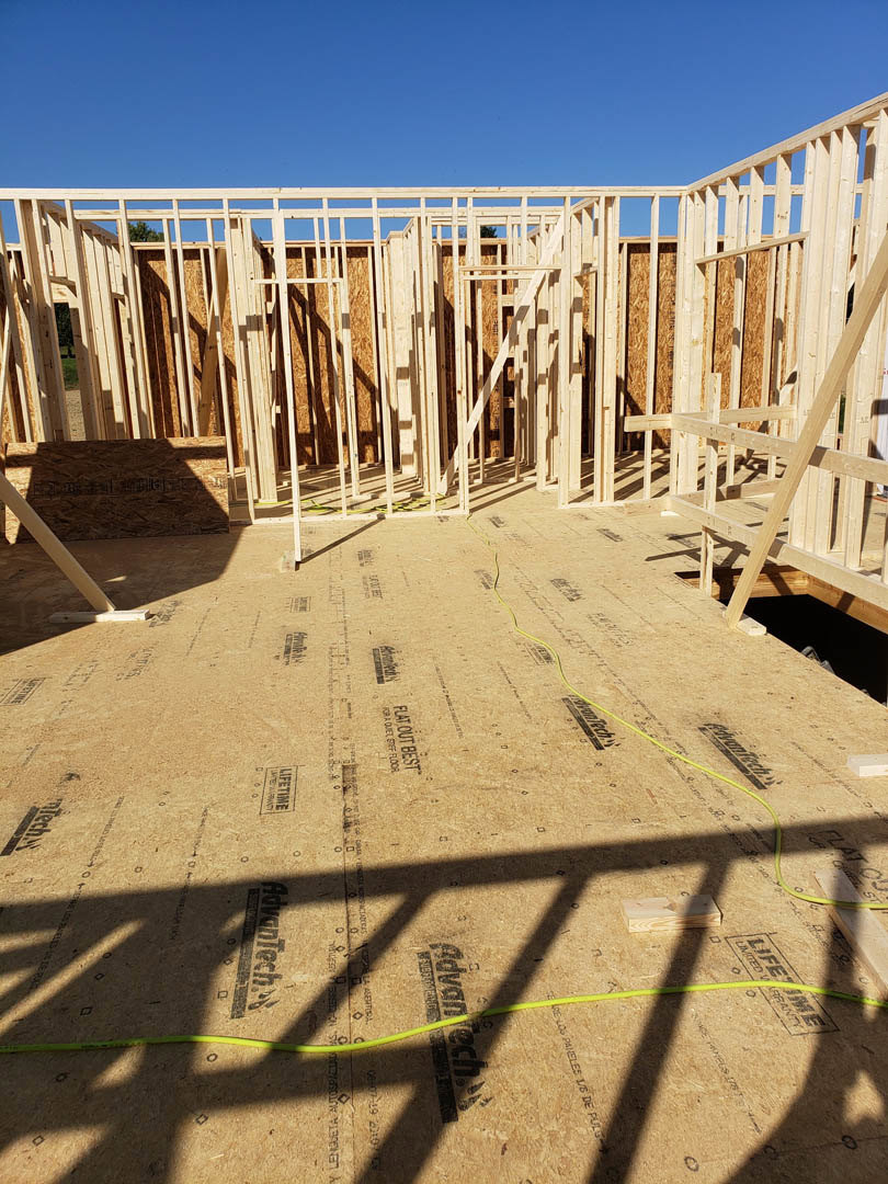 Exposed wooden framing and plywood sheathing of a house under construction, yellow rope stretched across the site, white railing in foreground, clear blue sky overhead