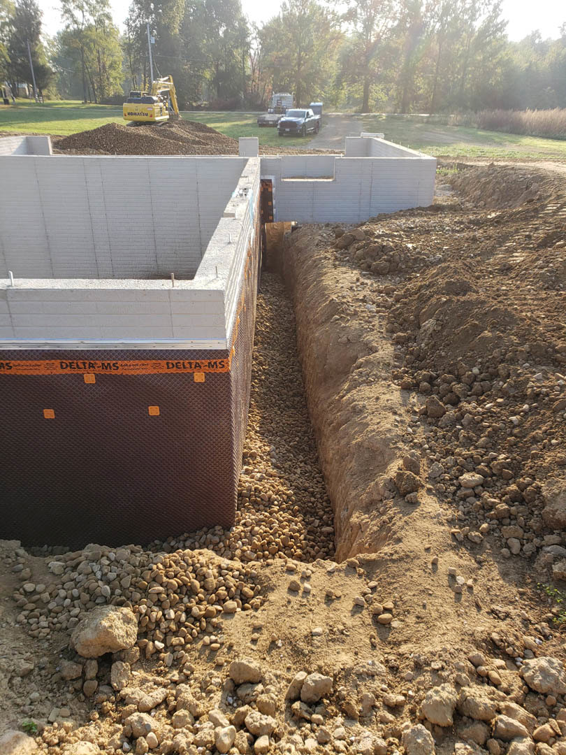 Construction site with exposed soil trench, scattered rocks, yellow backhoe, and partially built custom home exterior surrounded by trees