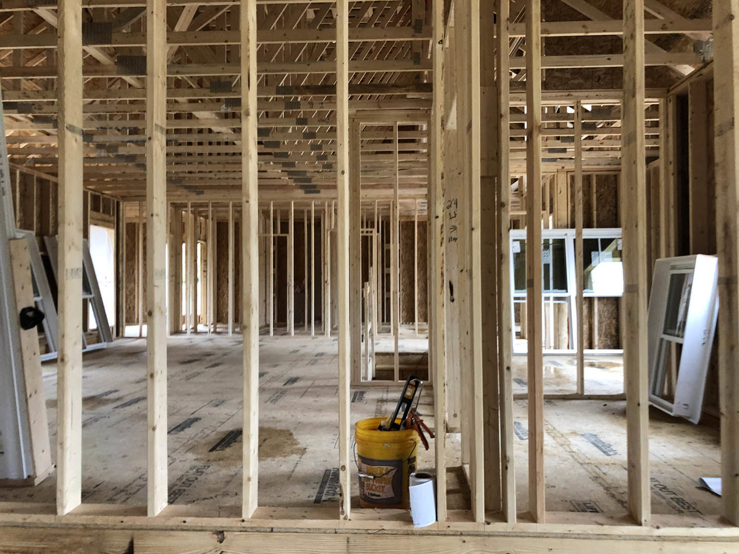 Framed interior under construction with exposed wooden beams, insulation panels, ladder, yellow bucket, roll of toilet paper, and hand holding a tool near scattered building