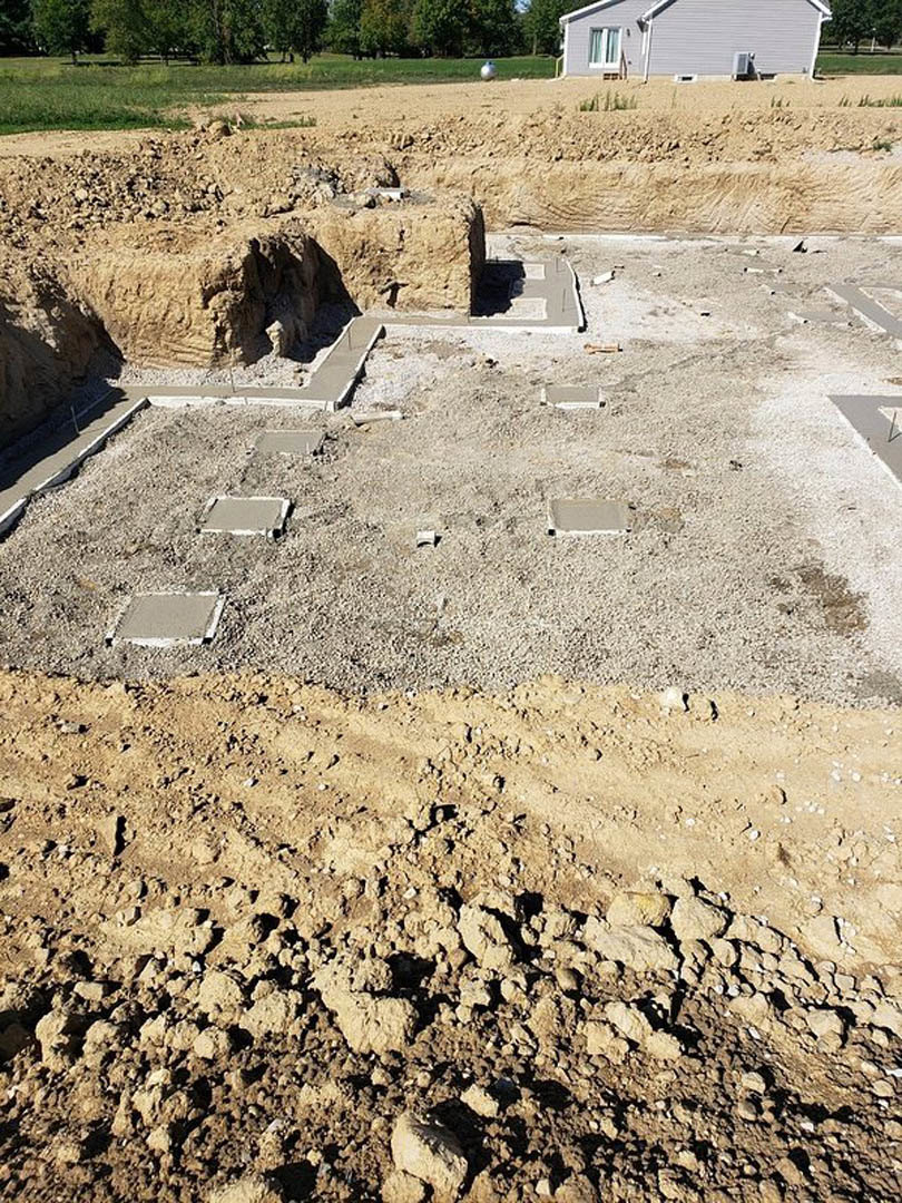 Concrete block foundation under construction with exposed soil, metal pole, and white wall; pile of dirt and scattered rocks visible on site.