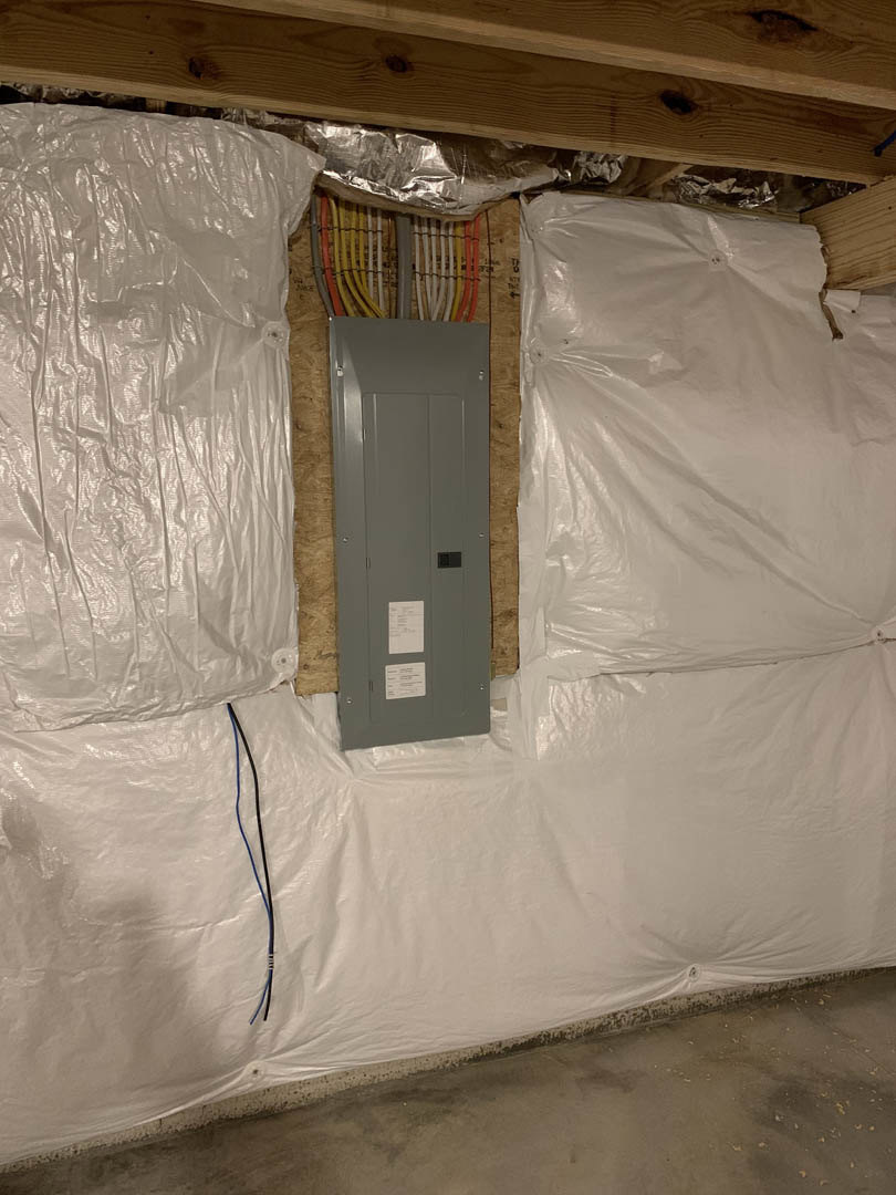 Grey electrical panel with bundled wires mounted on unfinished basement wall, exposed wooden beams, plastic sheeting, and concrete floor visible.