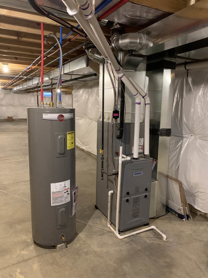 White water heater and cylindrical boiler installed against a wall, metal pipes connecting to ceiling and floor, surrounded by light-colored finishes in a utility room.