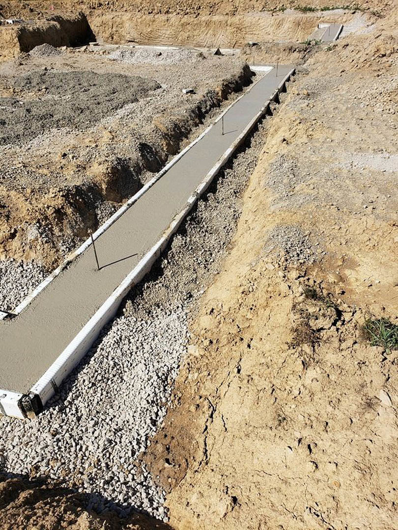 Concrete drainage channel set in bare dirt and gravel, bordered by a curb, with sparse vegetation nearby
