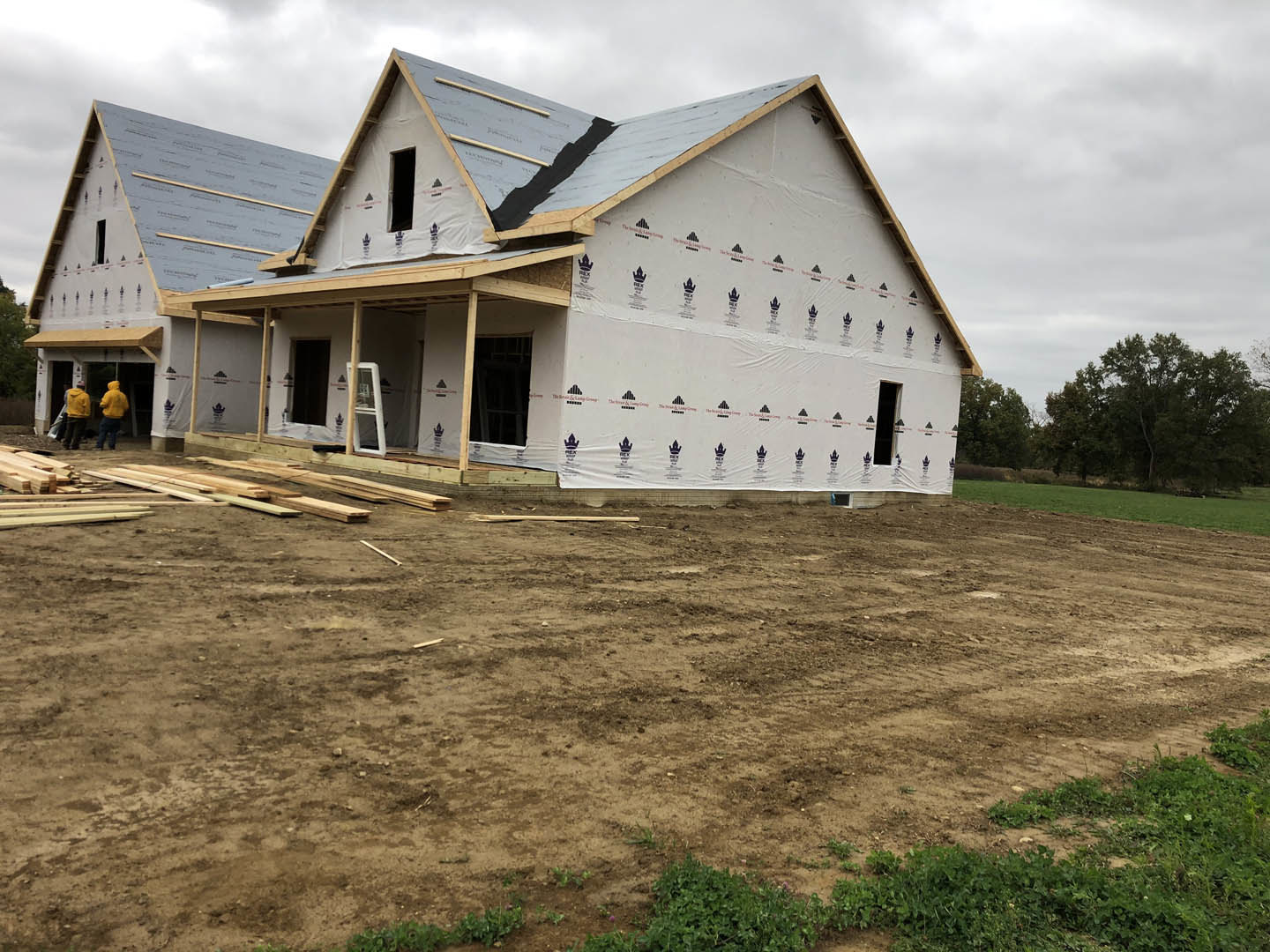 Partially built house with exposed framing and roof, surrounded by dirt field, construction materials, and several men in yellow jackets; trees visible in background under cloudy