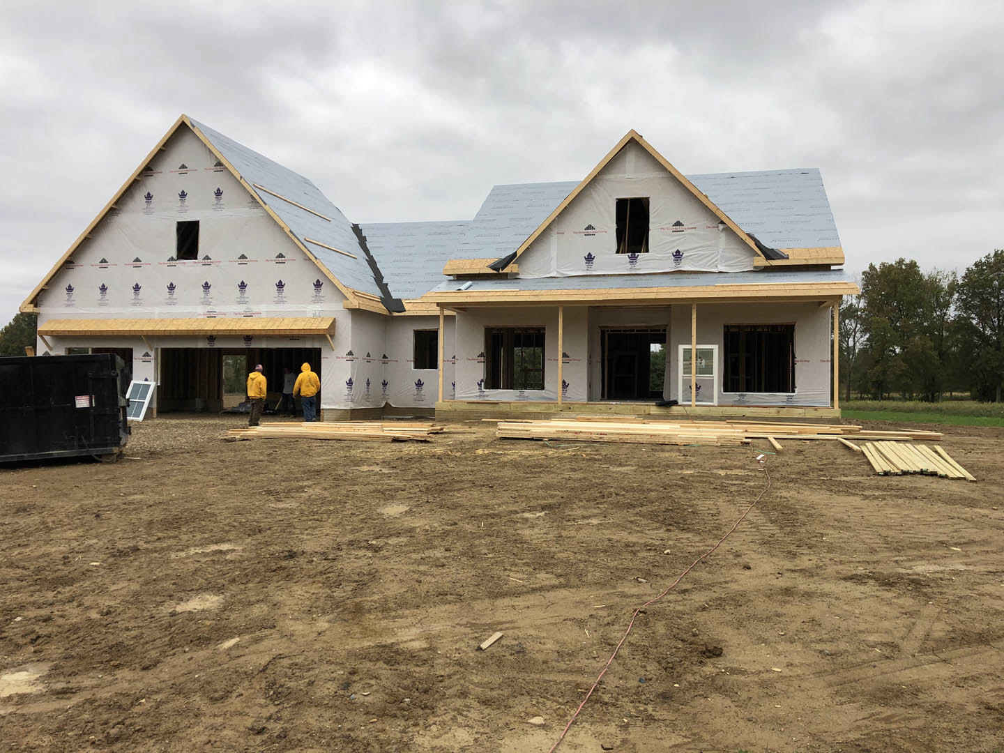 Two-story house under construction with exposed framing, dirt ground scattered with wood pieces, people in yellow jackets standing near the entrance, black container with white