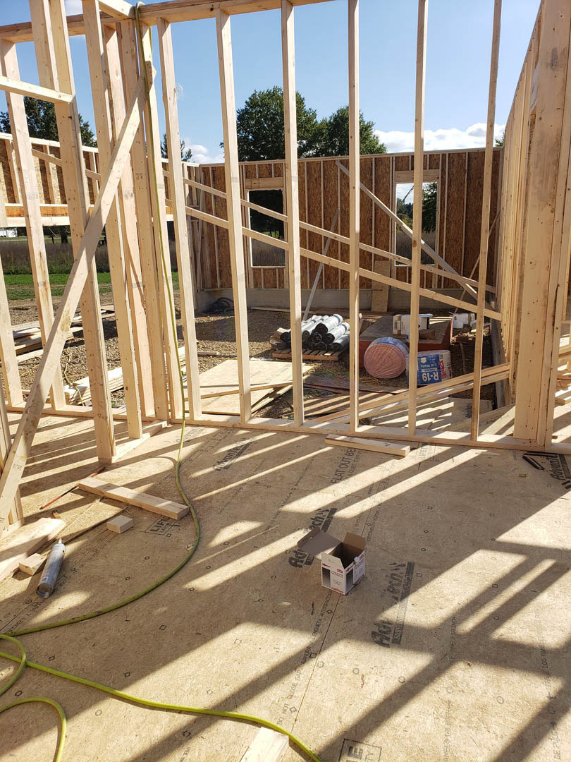 Wood-framed house under construction with exposed beams, plywood flooring, and scattered lumber; large blue-wrapped cylinder and cardboard box with white label on ground